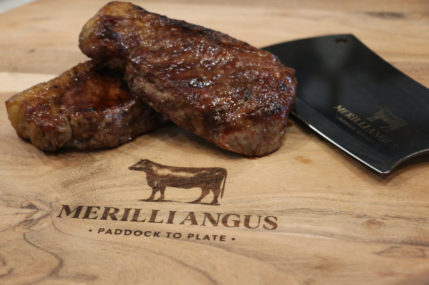 Cooked beef steaks on wooden board with cleaver, Merilli Angus paddock to plate logo